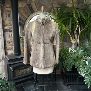 May include: A vintage fur coat in shades of brown and tan, displayed on a mannequin. The coat has a high collar and a fitted waist. The background includes a stone wall, a fireplace, and lush greenery, suggesting a rustic setting.