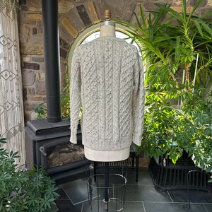 K&ouml;nnte beinhalten: Ein hellgrauer Zopfstrickpullover, der auf einer Schaufensterpuppe pr&auml;sentiert wird. Der Pullover hat ein strukturiertes Muster und lange &Auml;rmel. Der Hintergrund umfasst einen Kamin, Pflanzen und ein Fenster.