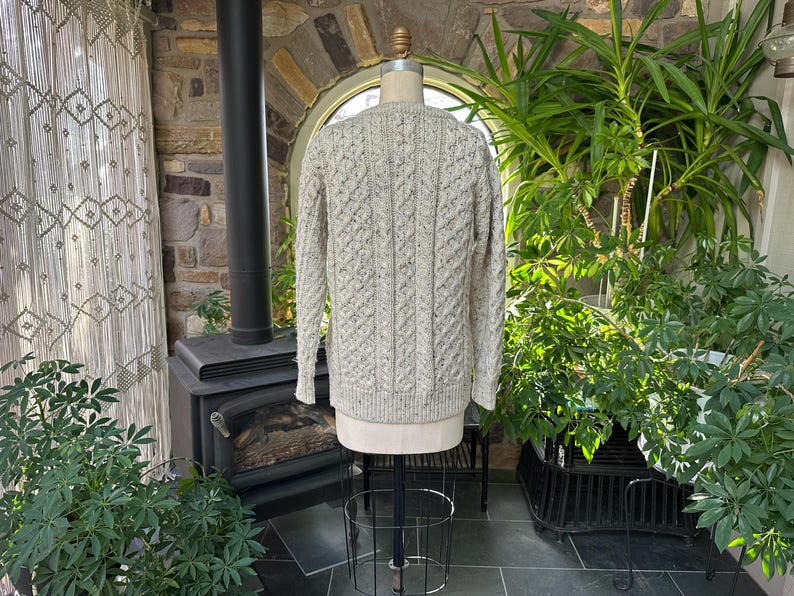 K&ouml;nnte beinhalten: Ein hellgrauer Zopfstrickpullover, der auf einer Schaufensterpuppe pr&auml;sentiert wird. Der Pullover hat ein strukturiertes Muster und lange &Auml;rmel. Der Hintergrund umfasst einen Kamin, Pflanzen und ein Fenster.