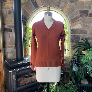 Puede incluir: Un suéter con cuello en V de color óxido exhibido en un maniquí. El suéter tiene puños y dobladillo acanalados. El maniquí está sobre un soporte negro, con el número "2" visible. El fondo incluye una pared de piedra, plantas y una ventana.