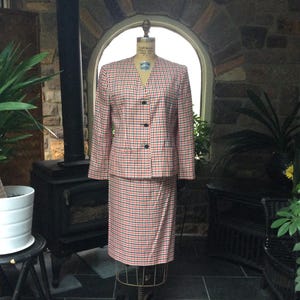 May include: A vintage two-piece suit displayed on a mannequin. The suit features a red, black, and white checkered pattern. The jacket has a V-neck and black buttons. The skirt is knee-length. The suit is displayed in front of a stone wall and a window.