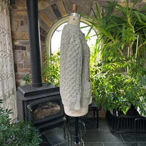 K&ouml;nnte beinhalten: Hellgraue Strickjacke mit Zopfmuster und zwei Vordertaschen, auf einer Schaufensterpuppe pr&auml;sentiert. Der Pullover hat ein strukturiertes Muster und besteht aus einem dicken, warmen Material. Der Pullover wird in einem Raum mit Kamin und Pflanzen pr&auml;sentiert.