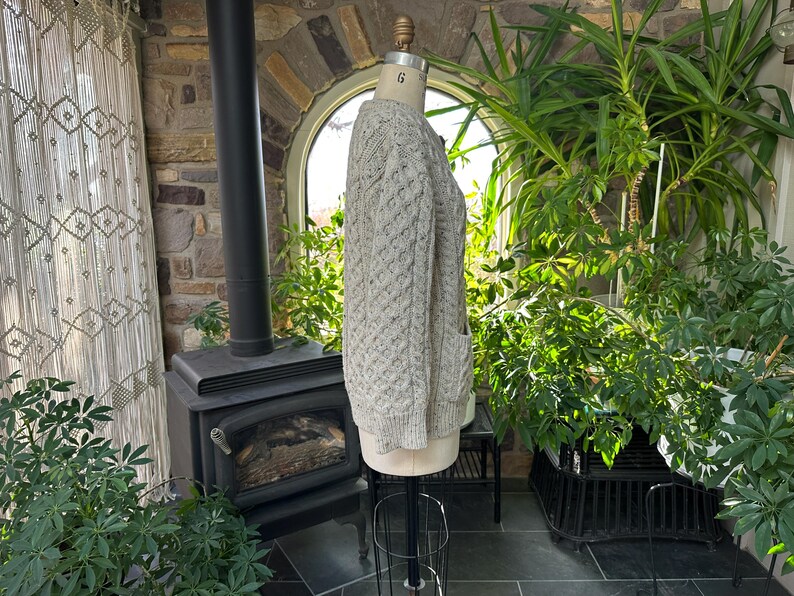 K&ouml;nnte beinhalten: Hellgraue Strickjacke mit Zopfmuster und zwei Vordertaschen, auf einer Schaufensterpuppe pr&auml;sentiert. Der Pullover hat ein strukturiertes Muster und besteht aus einem dicken, warmen Material. Der Pullover wird in einem Raum mit Kamin und Pflanzen pr&auml;sentiert.
