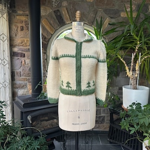 May include: Cream-colored knitted zip-up sweater with green trim and tree designs. The sweater is displayed on a mannequin. The background includes a brick wall, plants, and a fireplace.