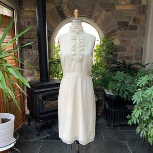 May include: Cream-colored sleeveless dress on a mannequin. The dress features a high, ruffled collar and a fitted waist. The fabric has a subtle dotted pattern. The dress is knee-length and appears to be vintage.