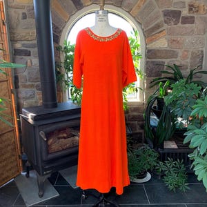 May include: A bright orange maxi dress with elbow-length sleeves and a jewel-trimmed neckline. The dress is displayed on a mannequin, highlighting its flowing shape. The dress is made of a soft, textured fabric.