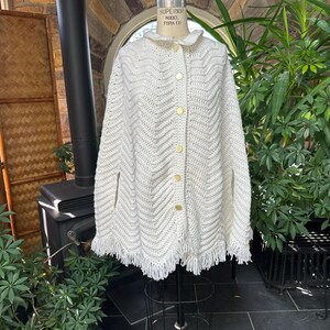 Op de afbeelding: Een crèmekleurige, gehaakte cape met een geschulpte kraag en franjes. De cape heeft een chevronpatroon en is vastgemaakt met goudkleurige knopen. Het kledingstuk wordt op een mannequin getoond.