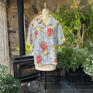 Może przedstawiać: Jasnoniebieska koszula hawajska z tropikalnym kwiatowym nadrukiem z czerwonych kwiat&oacute;w hibiskusa i ż&oacute;łtych ananas&oacute;w. Koszula z kr&oacute;tkim rękawem jest prezentowana na manekinie. Koszula ma zapięcie na guziki z przodu i kołnierz.