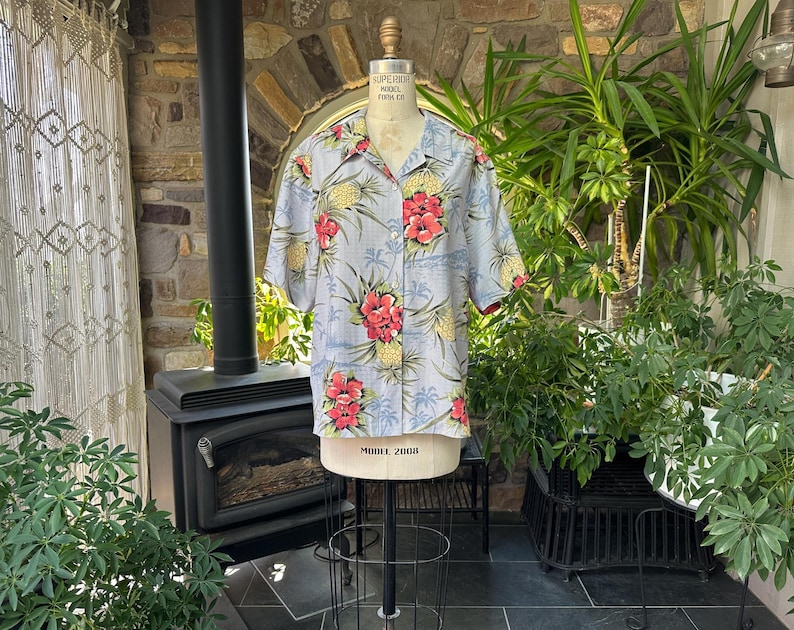 Może przedstawiać: Jasnoniebieska koszula hawajska z tropikalnym kwiatowym nadrukiem z czerwonych kwiat&oacute;w hibiskusa i ż&oacute;łtych ananas&oacute;w. Koszula z kr&oacute;tkim rękawem jest prezentowana na manekinie. Koszula ma zapięcie na guziki z przodu i kołnierz.