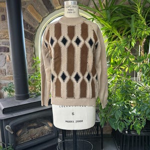 May include: A long-sleeve sweater featuring a beige, brown, and black diamond argyle pattern on the front. The garment has a ribbed mock neck collar, ribbed cuffs, and a ribbed hem. The sweater is displayed on a mannequin with visible text.