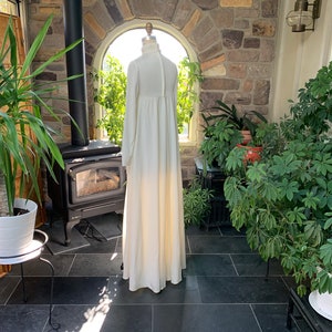Peut inclure: Une longue robe ivoire &agrave; manches longues et col montant est pr&eacute;sent&eacute;e sur un mannequin. La robe a une silhouette fluide et une fermeture boutonn&eacute;e dans le dos. Le cadre est une v&eacute;randa avec des plantes et un mur en pierre.