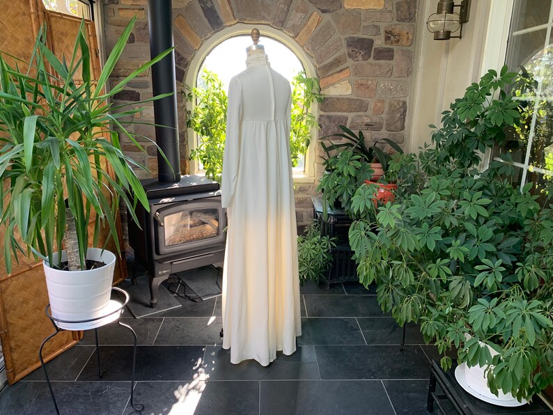 Peut inclure: Une longue robe ivoire &agrave; manches longues et col montant est pr&eacute;sent&eacute;e sur un mannequin. La robe a une silhouette fluide et une fermeture boutonn&eacute;e dans le dos. Le cadre est une v&eacute;randa avec des plantes et un mur en pierre.