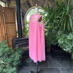 Puede incluir: Un camis&oacute;n rosa vintage en un maniqu&iacute;. El camis&oacute;n tiene un dise&ntilde;o transparente y fluido con un escote y dobladillo con ribete de encaje. El maniqu&iacute; est&aacute; sobre un soporte negro, y el fondo incluye plantas y una chimenea.