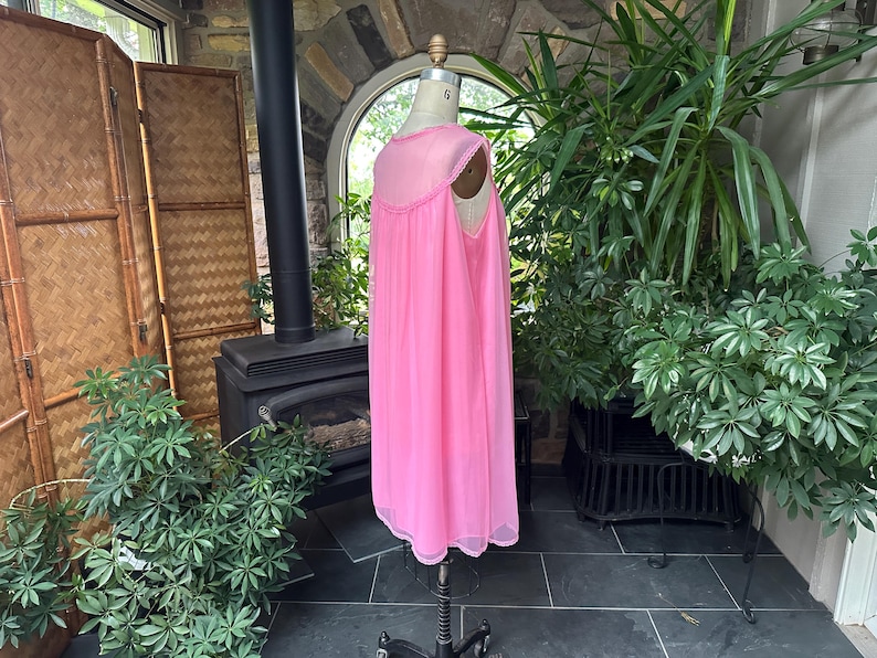 Puede incluir: Un camis&oacute;n rosa vintage en un maniqu&iacute;. El camis&oacute;n tiene un dise&ntilde;o transparente y fluido con un escote y dobladillo con ribete de encaje. El maniqu&iacute; est&aacute; sobre un soporte negro, y el fondo incluye plantas y una chimenea.