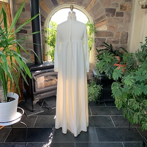 Peut inclure: Une longue robe ivoire &agrave; manches longues et col montant. La robe a un corsage ajust&eacute; et une jupe fluide. Elle est pr&eacute;sent&eacute;e sur un mannequin dans une pi&egrave;ce ensoleill&eacute;e avec des plantes et une chemin&eacute;e.