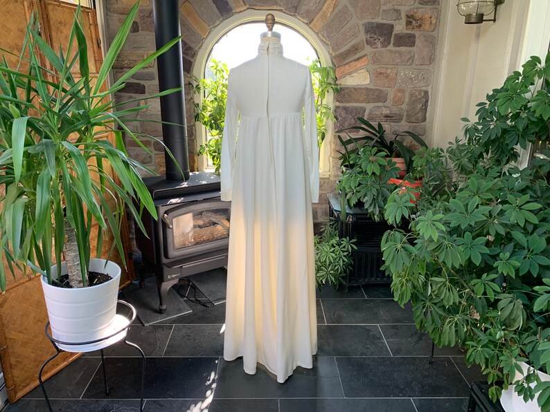 Peut inclure: Une longue robe ivoire &agrave; manches longues et col montant. La robe a un corsage ajust&eacute; et une jupe fluide. Elle est pr&eacute;sent&eacute;e sur un mannequin dans une pi&egrave;ce ensoleill&eacute;e avec des plantes et une chemin&eacute;e.