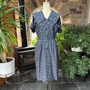 May include: A vintage blue dress with a white geometric pattern, displayed on a dress form. The dress features a large collar, short sleeves, and a button-down front. The dress is knee-length and appears to be made of a textured fabric.