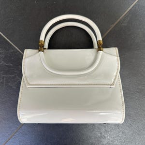 May include: A glossy white handbag with a structured rectangular body and a curved top flap. The bag features a single rounded handle attached with gold-toned metal hardware accents at the base of the handle on both sides of the bag.