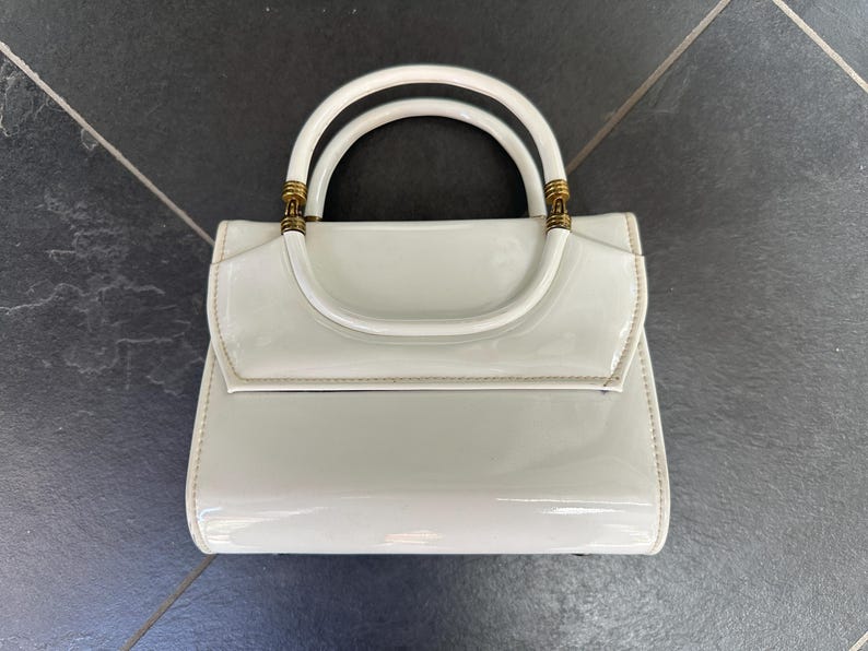 May include: A glossy white handbag with a structured rectangular body and a curved top flap. The bag features a single rounded handle attached with gold-toned metal hardware accents at the base of the handle on both sides of the bag.