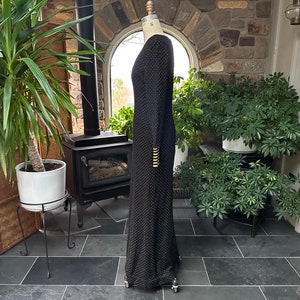 Pu&ograve; includere: Un abito nero lungo fino al pavimento con un sottile motivo a pois. L'abito a maniche lunghe presenta bottoni color oro sui polsini. L'abito &egrave; esposto su un manichino in un ambiente interno ben illuminato.