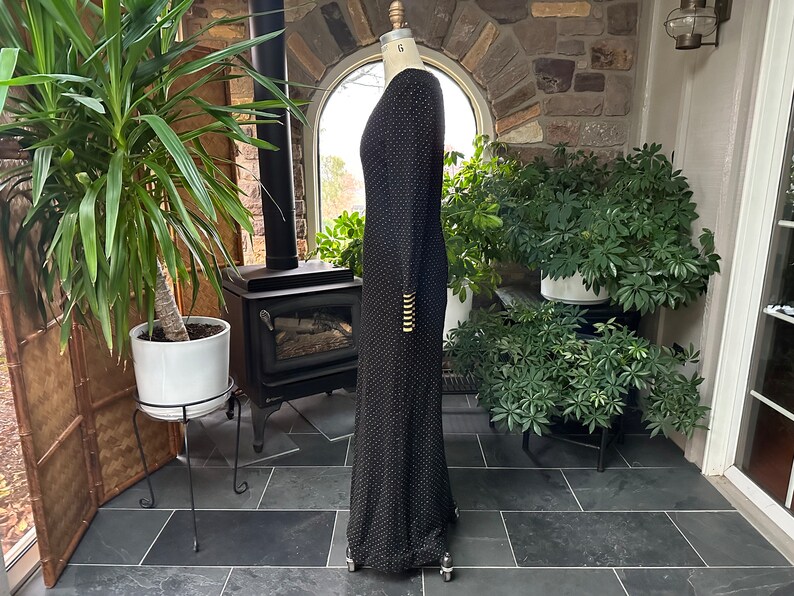 Pu&ograve; includere: Un abito nero lungo fino al pavimento con un sottile motivo a pois. L'abito a maniche lunghe presenta bottoni color oro sui polsini. L'abito &egrave; esposto su un manichino in un ambiente interno ben illuminato.