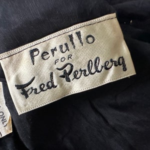 Pu&ograve; includere: Primo piano di un capo nero con un'etichetta che recita "Perullo for Fred Perlberg" in corsivo nero. Un'etichetta pi&ugrave; piccola sotto recita "Dry Clean Only". Il tessuto sembra essere un materiale scuro, possibilmente vintage.