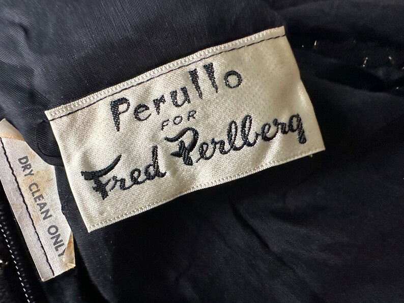 Pu&ograve; includere: Primo piano di un capo nero con un'etichetta che recita "Perullo for Fred Perlberg" in corsivo nero. Un'etichetta pi&ugrave; piccola sotto recita "Dry Clean Only". Il tessuto sembra essere un materiale scuro, possibilmente vintage.