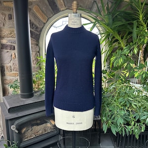 Puede incluir: Un jersey de cuello alto azul marino exhibido en un maniquí. El jersey de manga larga está hecho de un material suave y tiene dobladillo y puños acanalados. El maniquí está sobre un soporte metálico, con plantas y una pared de piedra en el fondo.