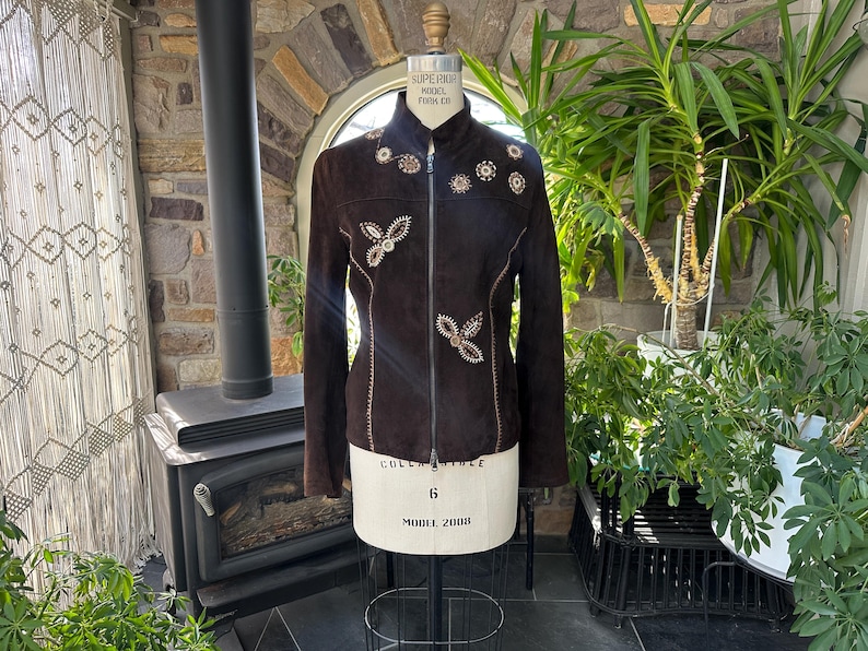 May include: A dark brown suede jacket with a high collar and a full-length zipper. The jacket is adorned with embroidered floral and butterfly designs in cream and gold. The jacket is displayed on a mannequin.