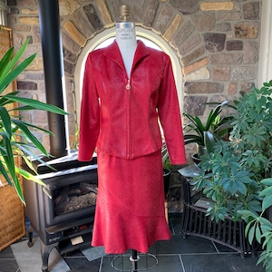 May include: A red two-piece suit with a textured pattern, displayed on a mannequin. The jacket has a zip-up front and collar, and the skirt has a flared hem. The suit is in a room with plants and a fireplace, creating a stylish display.