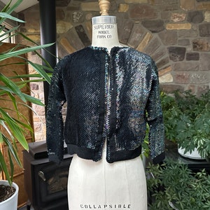 May include: A black sequined jacket displayed on a mannequin. The jacket has a round neckline, long sleeves, and a zipper closure. The sequins create an iridescent effect, reflecting multiple colors. The jacket is a cropped style with a ribbed hem and cuffs.