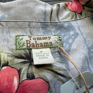 Może przedstawiać: Jasnoniebieska jedwabna koszula Tommy Bahama z kwiatowym wzorem w kolorze czerwonym, r&oacute;żowym i zielonym. Koszula ma metkę z napisem "100% Silk Made in China". Agrafka mocuje metkę do koszuli.