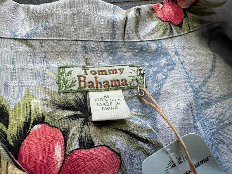 Może przedstawiać: Jasnoniebieska jedwabna koszula Tommy Bahama z kwiatowym wzorem w kolorze czerwonym, r&oacute;żowym i zielonym. Koszula ma metkę z napisem "100% Silk Made in China". Agrafka mocuje metkę do koszuli.