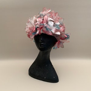 May include: A floral hat with pink, white, and blue fabric flowers. The hat is displayed on a black mannequin head. The flowers are layered and create a full, textured look, perfect for a special occasion.