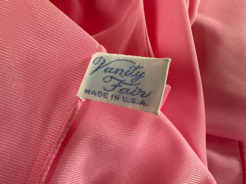 Puede incluir: Primer plano de una tela rosa con una etiqueta que dice "Vanity Fair MADE IN U.S.A." La tela tiene una textura suave y sedosa, posiblemente una prenda. La etiqueta es rectangular con texto azul.