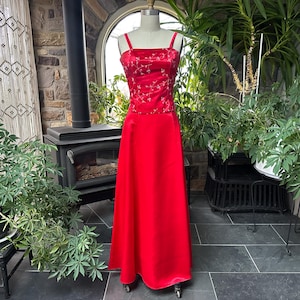 Op de afbeelding: Een rode galajurk op een mannequin. Het lijfje is donkerder rood met een bloemmotief en de rok is effen, vloeiend rood. De jurk heeft spaghettibandjes en een lange rok. De jurk wordt getoond in een kamer met planten en een open haard.