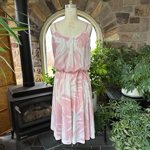 Peut inclure: Une robe sans manches avec un motif de feuilles tropicales rose clair et blanc, une encolure dégagée avec de fines bretelles, et une taille élastiquée avec une ceinture à nouer assortie, présentée sur un mannequin de couture.