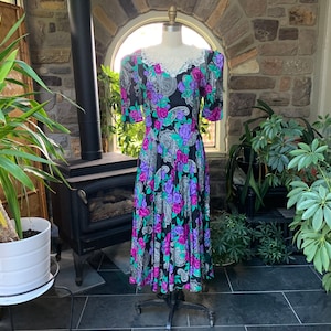 May include: A black midi dress featuring a vibrant floral and paisley pattern in shades of magenta, purple, and teal. The dress has short sleeves, a defined waist, and a white lace collar detail at the neckline, displayed on a dress form mannequin.