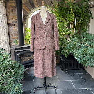 Könnte beinhalten: Ein zweiteiliger Vintage-Anzug aus mauvefarbenem Tweed-Stoff. Der Anzug besteht aus einem Blazer mit Zwei-Knopf-Verschluss und einem passenden knielangen Rock. Der Anzug wird auf einer Schaufensterpuppe präsentiert, die sein klassisches Design und seine maßgeschneiderte Passform hervorhebt.