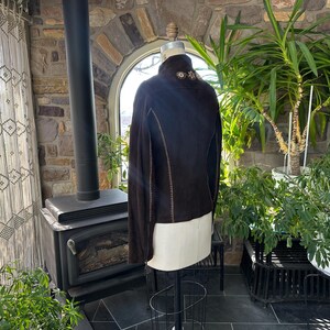 May include: A dark brown suede jacket with a high collar and decorative stitching. The jacket is displayed on a mannequin, showcasing its tailored fit and design. The collar features embroidered detailing. The jacket is a stylish outerwear piece.