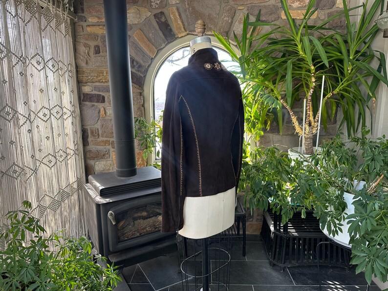 May include: A dark brown suede jacket with a high collar and decorative stitching. The jacket is displayed on a mannequin, showcasing its tailored fit and design. The collar features embroidered detailing. The jacket is a stylish outerwear piece.