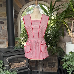 Op de afbeelding: Een rood-wit gestreept schort met een vierkante halslijn, brede schouderbanden en twee voorzakken. Het schort is afgewerkt met rode biezen en witte kanten details langs het lijfje en de randen van de zakken, gepresenteerd op een paspop.