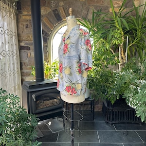 Może przedstawiać: Jasnoniebieska koszula hawajska z kwiatowym i ananasowym wzorem. Koszula ma kr&oacute;tkie rękawy z czerwonym wykończeniem i jest prezentowana na manekinie. Tło obejmuje rośliny, kominek i okno.