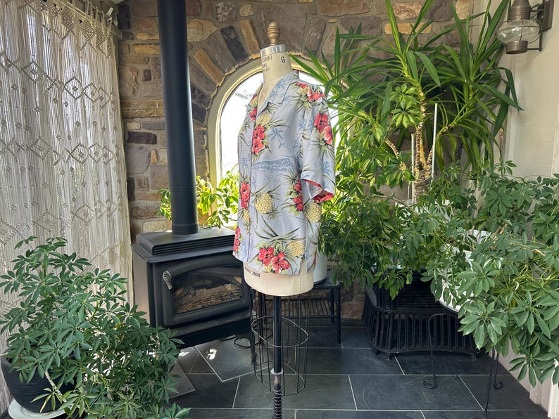 Może przedstawiać: Jasnoniebieska koszula hawajska z kwiatowym i ananasowym wzorem. Koszula ma kr&oacute;tkie rękawy z czerwonym wykończeniem i jest prezentowana na manekinie. Tło obejmuje rośliny, kominek i okno.