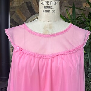 Puede incluir: Un camis&oacute;n rosa vintage con canes&uacute; transparente y ribete con volantes. El camis&oacute;n presenta un delicado lazo rosa en el escote y se exhibe en un maniqu&iacute;. La prenda est&aacute; hecha de una tela ligera y fluida.