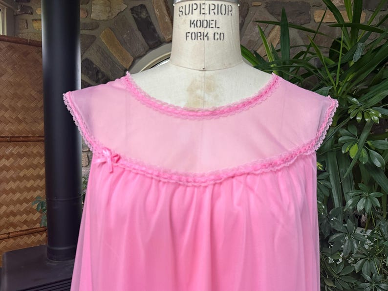 Puede incluir: Un camis&oacute;n rosa vintage con canes&uacute; transparente y ribete con volantes. El camis&oacute;n presenta un delicado lazo rosa en el escote y se exhibe en un maniqu&iacute;. La prenda est&aacute; hecha de una tela ligera y fluida.