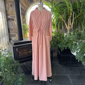 Könnte beinhalten: Ein langer, hellrosa Bademantel mit Reißverschluss vorne, langen Ärmeln und zwei Vordertaschen. Der Bademantel hat eine Kordelzug-Taille und wird auf einer Schaufensterpuppe präsentiert. Der Hintergrund umfasst einen Kamin, Pflanzen und ein Fenster.