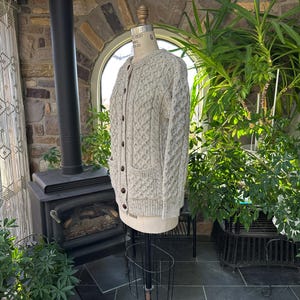 K&ouml;nnte beinhalten: Hellbeige Strickjacke mit Zopfmuster, dunklen braunen Kn&ouml;pfen und zwei Vordertaschen. Der Pullover wird auf einer Schaufensterpuppe pr&auml;sentiert. Der Hintergrund umfasst eine Steinmauer, einen schwarzen Ofen und verschiedene gr&uuml;ne Pflanzen.