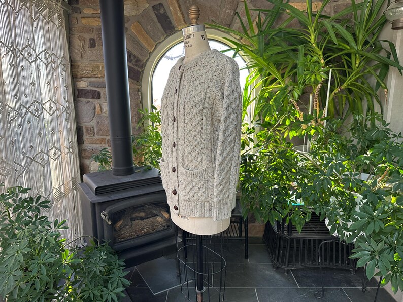 K&ouml;nnte beinhalten: Hellbeige Strickjacke mit Zopfmuster, dunklen braunen Kn&ouml;pfen und zwei Vordertaschen. Der Pullover wird auf einer Schaufensterpuppe pr&auml;sentiert. Der Hintergrund umfasst eine Steinmauer, einen schwarzen Ofen und verschiedene gr&uuml;ne Pflanzen.