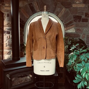 May include: A brown suede blazer with a notched lapel and a single-button closure. The jacket is displayed on a dress form. The blazer has two front pockets and a tailored fit. The background includes a stone wall and a window.